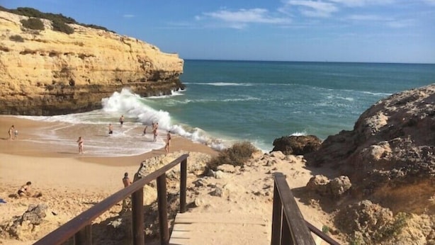 Una playa con un paseo marítimo de madera que lleva a la orilla, acantilados rocosos y el océano.