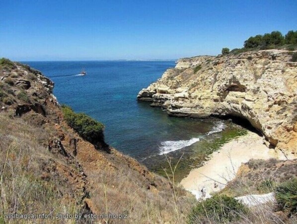 A coastal landscape with rocky cliffs, clear blue sea, and a sandy beach.