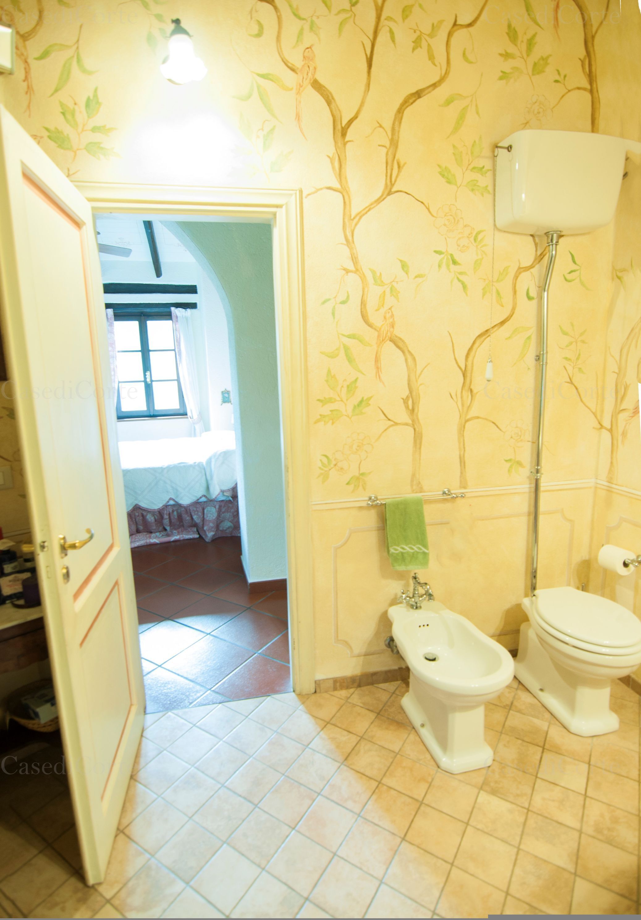 A bathroom with a toilet, bidet, and sink. There is a towel hanging on the wall and a window in the background.