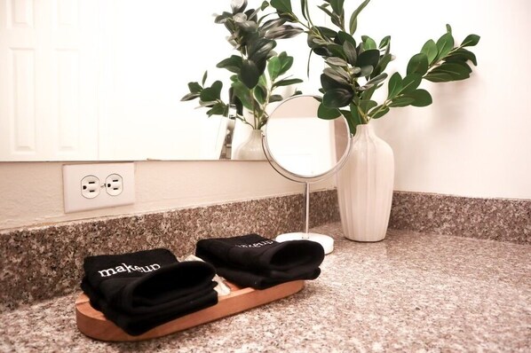 Un tocador de baño con encimera de granito, un jarrón blanco con follaje verde, un espejo redondo y un par de pantuflas negras con la palabra 'makeup' escrita.
