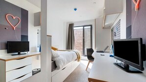A modern hotel room with a bed, a desk with a computer, a TV, and a decorative heart on the wall. - For Student Only Magdalen Street at Colchester - Campus Accommodation (Colchester)