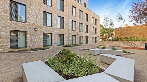 A modern building with a brick facade, multiple windows, and a courtyard with planters and seating areas. - For Student Only Magdalen Street at Colchester - Campus Accommodation (Colchester)