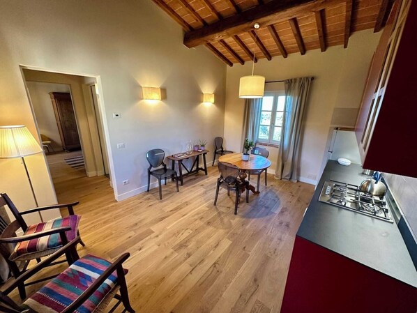 A cozy dining area with a wooden table and chairs, a kitchen with a gas stove, and a window with curtains. - Belvedere 19 (Bagno A Ripoli)