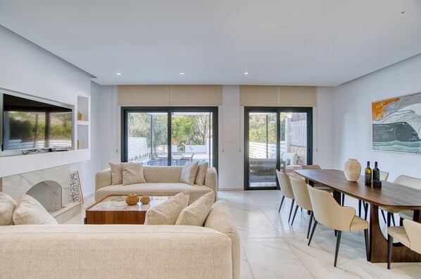 A modern living room with a sofa, coffee table, and dining area with chairs. There is a TV mounted on the wall and a painting above the dining table. - Magnus Villa - Three Bedroom Villa, Sleeps 6 (Tala)