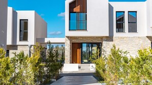A modern two-story house with a prominent wooden slatted window, a large glass door, and a stone facade. - Avalon Gardens Villa 21 (Emba)