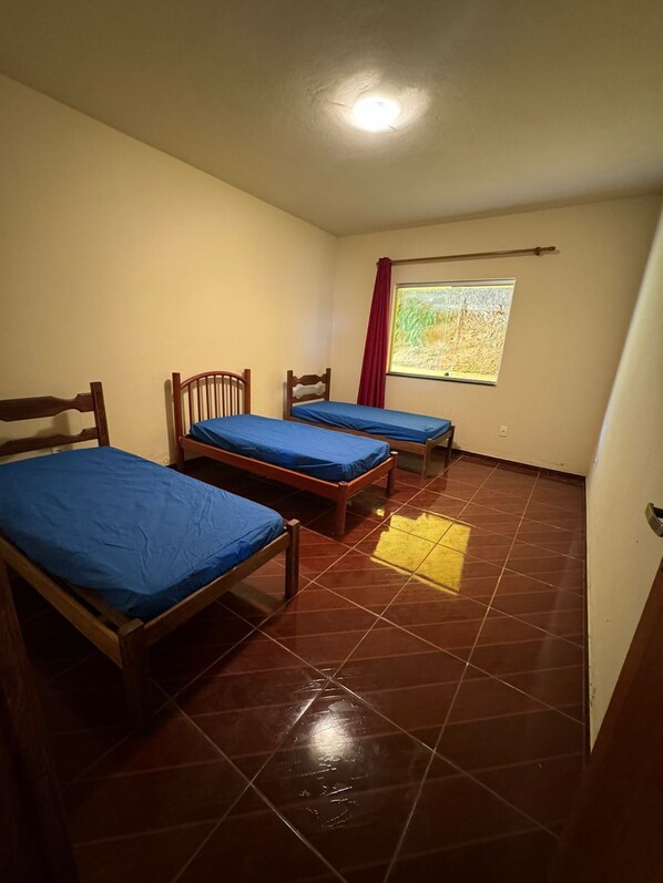Quarto com três camas de madeira, roupa de cama azul, uma janela com vista para um campo e uma cortina vermelha.