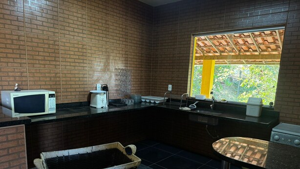 A kitchen with a microwave, toaster, sink, and a window with a view of greenery. - Sítio Sonho Real (Taquaraçu de Minas)