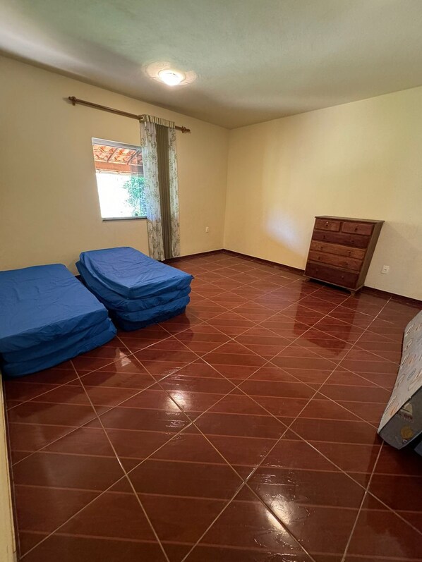 A room with a tiled floor, a window with curtains, a wooden dresser, and two beds covered with blue sheets. - Sítio Sonho Real (Taquaraçu de Minas)