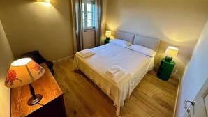 A bedroom with a large bed, wooden flooring, two bedside lamps, a nightstand, and a window with curtains. - Belvedere 18 (Bagno A Ripoli)