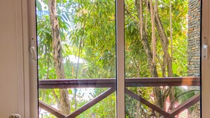 Ein Zimmer mit großem Fenster und Blick auf einen Garten mit Bäumen und einer Steinmauer.