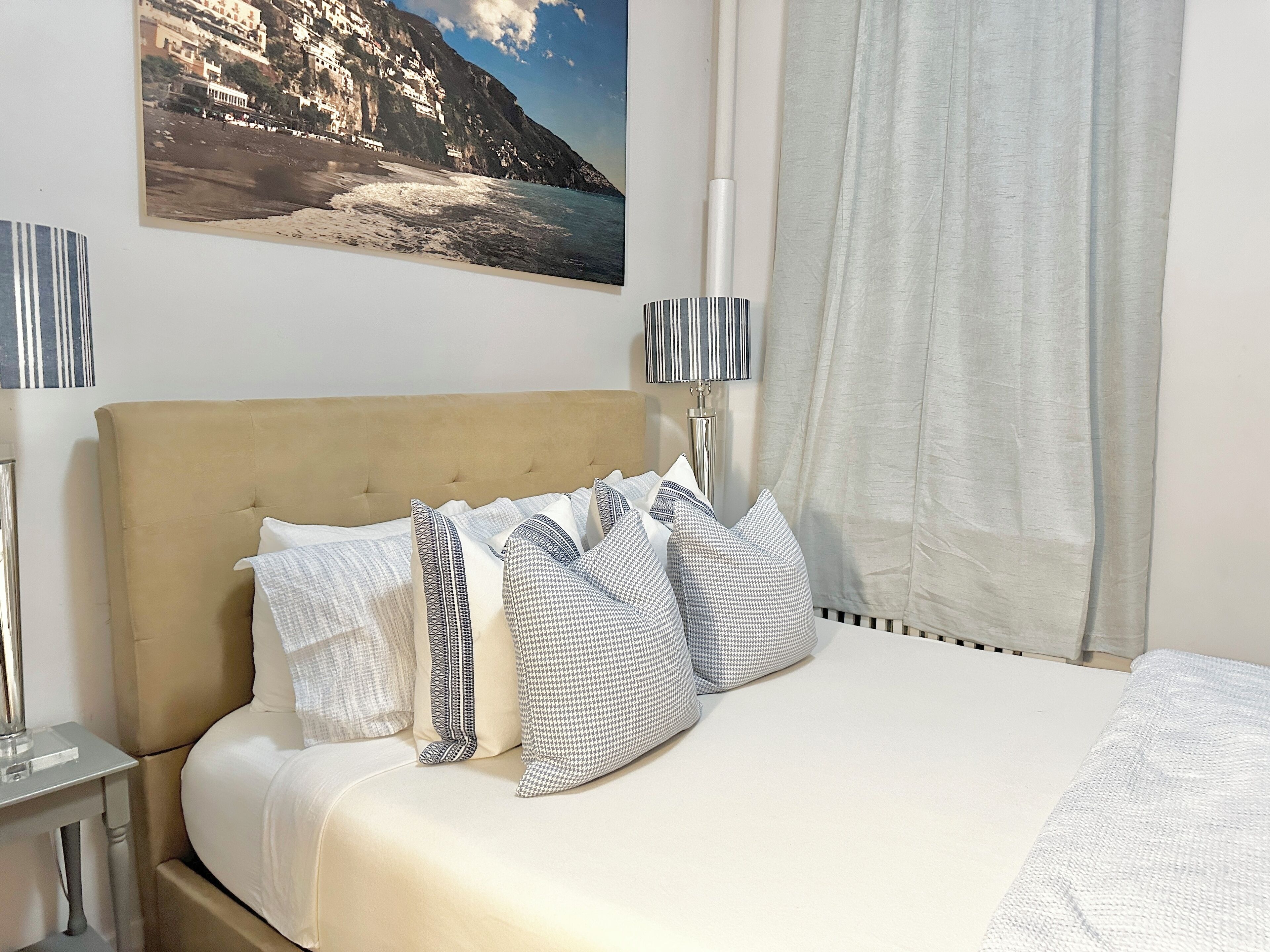 Una cama bien hecha con un cabecero color beige, sábanas blancas y almohadas estampadas. Encima de la cama, en la pared, hay un cuadro de un paisaje costero.