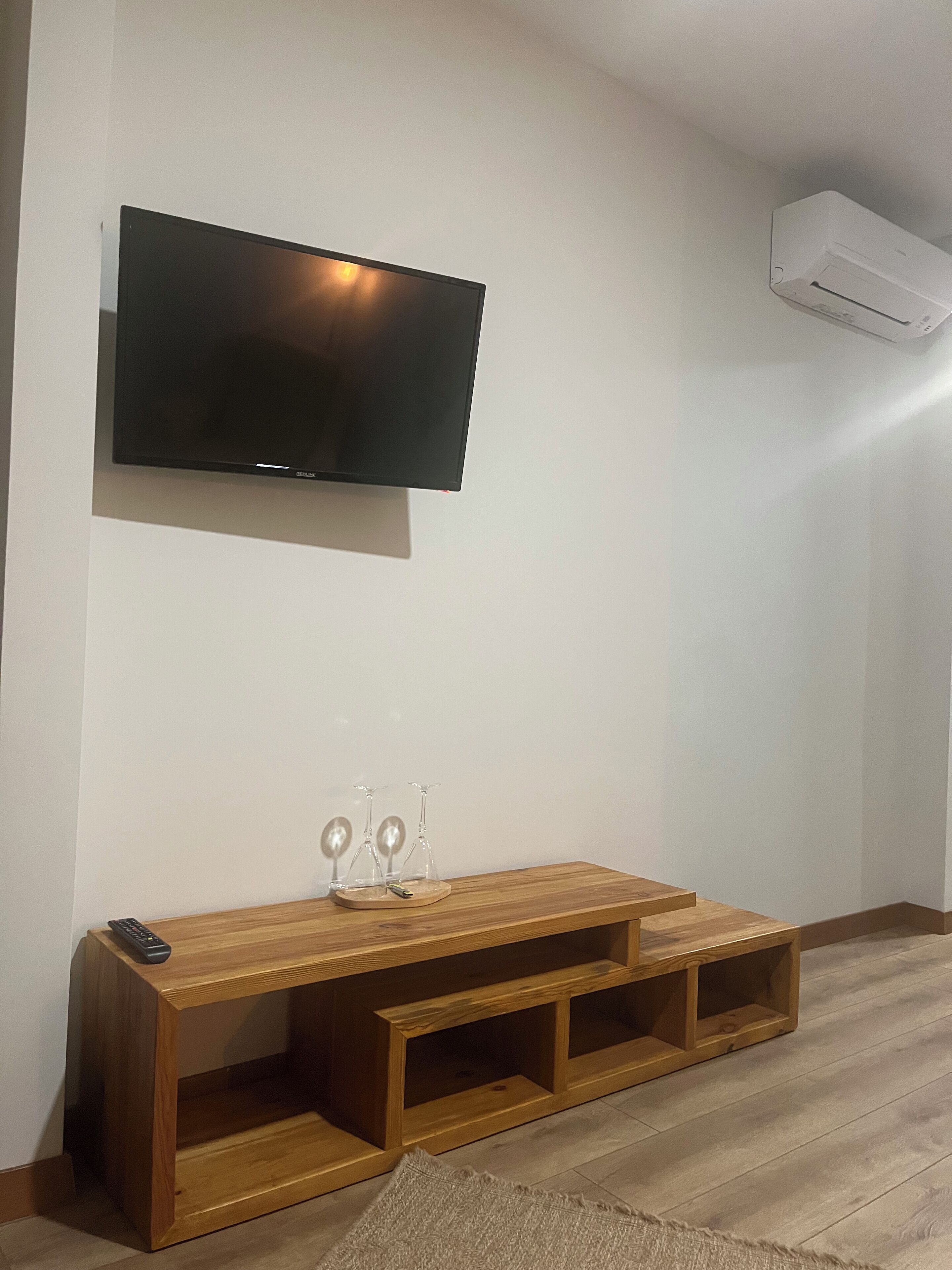 Uma sala de estar com televisão de tela plana fixada na parede, um rack de madeira com copos e um aparelho de ar condicionado.