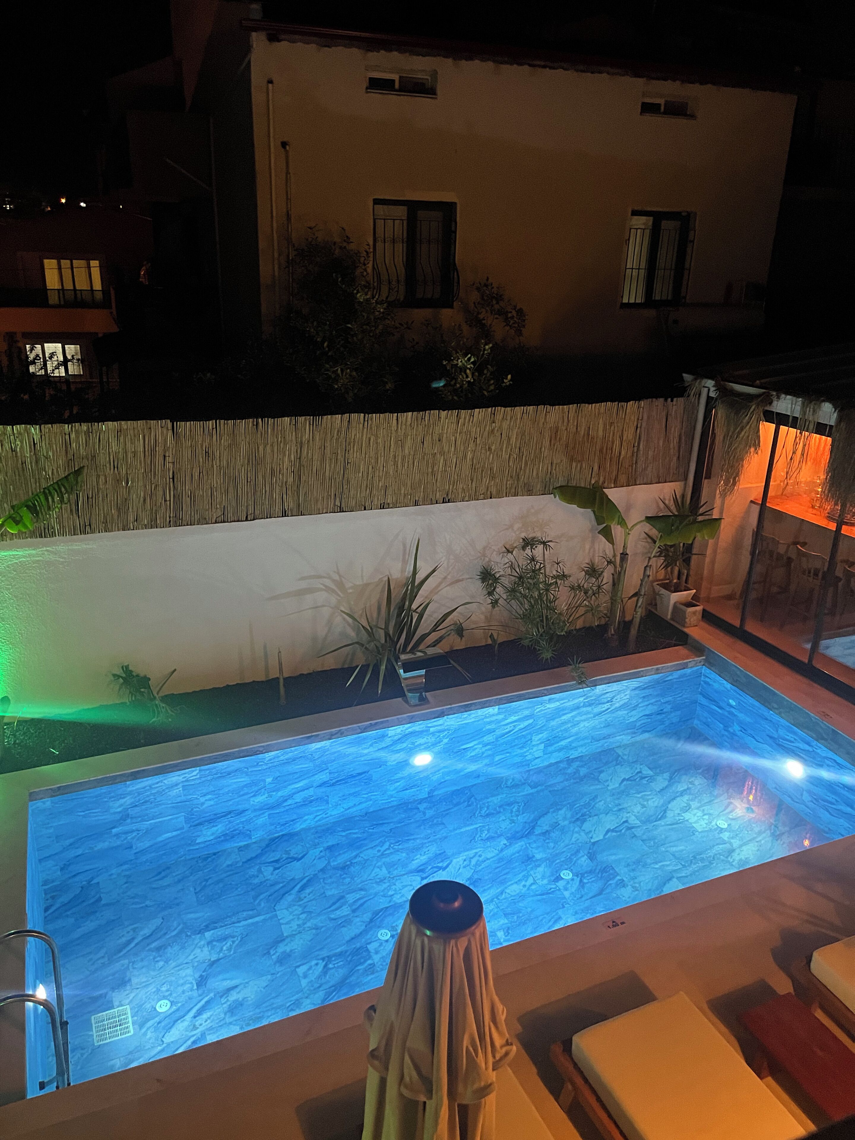 Piscina ao ar livre com cerca de bambu, cadeiras de descanso e área de estar coberta.