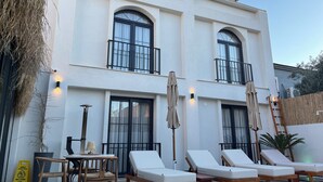 A two-story building with a white exterior, large windows, and balconies. There are lounge chairs and a table with a lamp, suggesting a patio area. - Güzergah Urla (Urla)