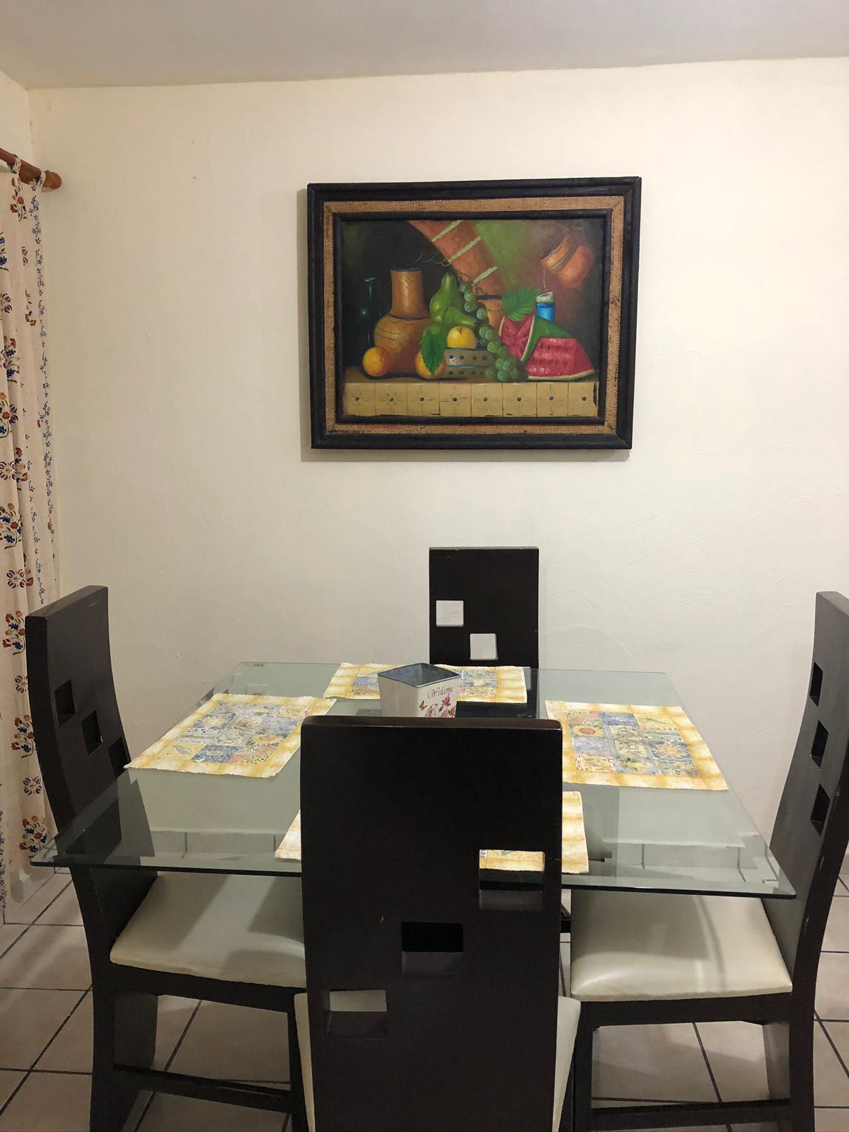 A dining area with a glass table, black chairs, and a framed painting above.