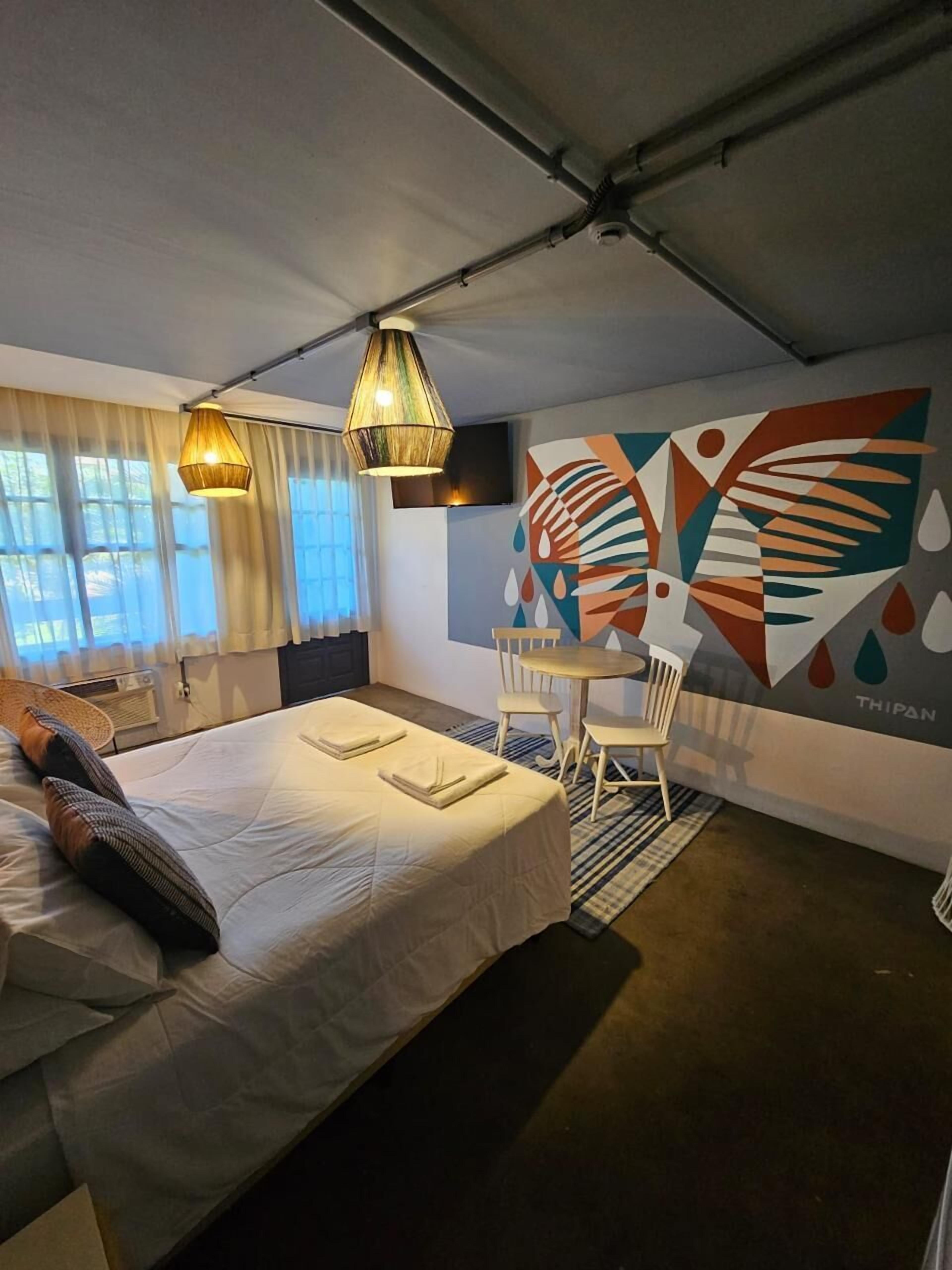 A hotel room with a bed, a small table, and a chair. There is a window with curtains, a wall with a colorful abstract design, and a ceiling with exposed beams.