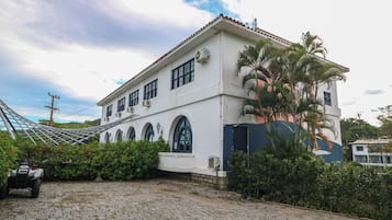 A white building with arched windows and a tiled roof, surrounded by greenery and a cobblestone path.