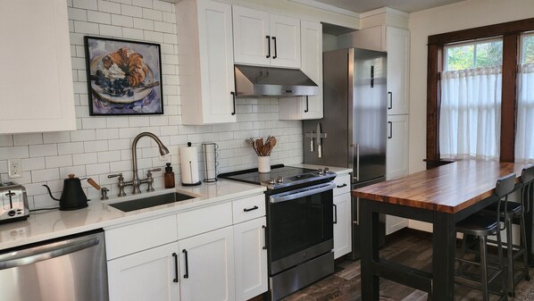 A modern kitchen with white cabinets, stainless steel appliances, a wooden dining table, and a window with curtains. - 2nd Floor Creekside Escape Near Ferries to Seattle (Port Orchard)