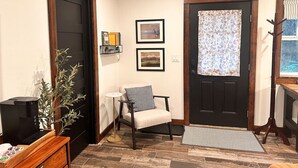 A living room with a black door, a white chair, a small table, a plant, and a television on a wooden stand. - Charming Creekside Studio Near Ferries to Seattle (Port Orchard)