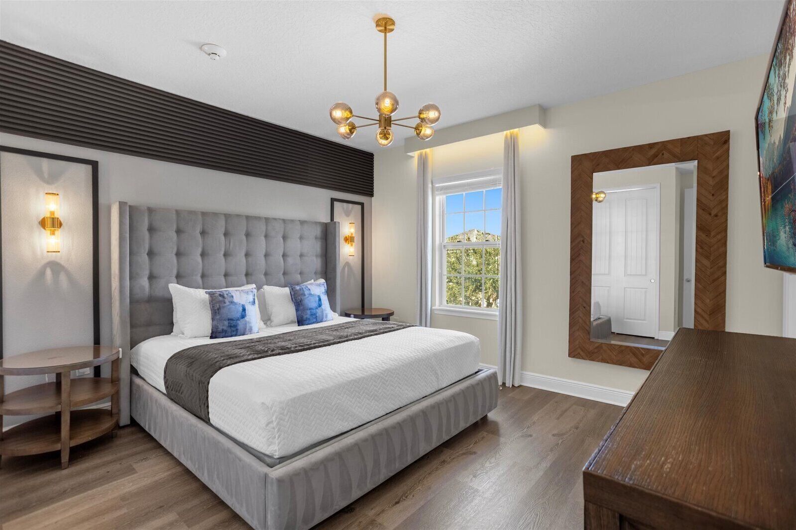 Un dormitorio moderno con un cabecero capitonado, una cama grande con ropa blanca y almohadas azules, una mesita de noche redonda de madera, un banco de madera y un espejo grande en la pared.