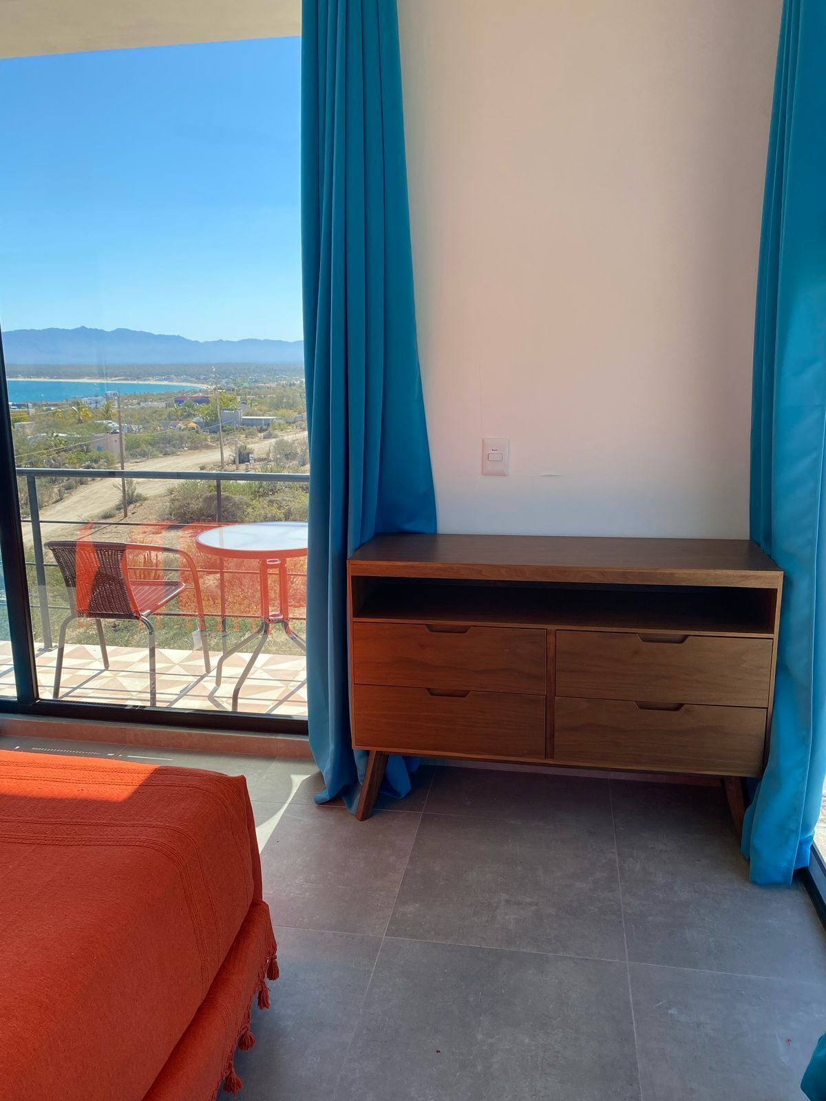 Een balkon met uitzicht op een strand en bergen, een houten bureau en een bed met een oranje deken.