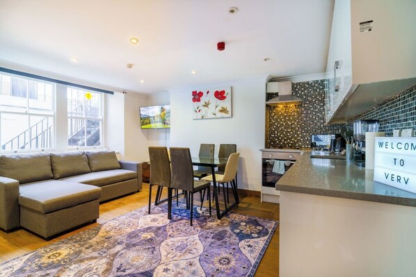A modern living room with a sofa, dining table, and chairs, a kitchen area with a tiled backsplash, and a welcome sign. - Angel x Kings Cross Rooms (London)