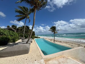 A poolside area with a clear view of the ocean, surrounded by palm trees and a sandy beach.
