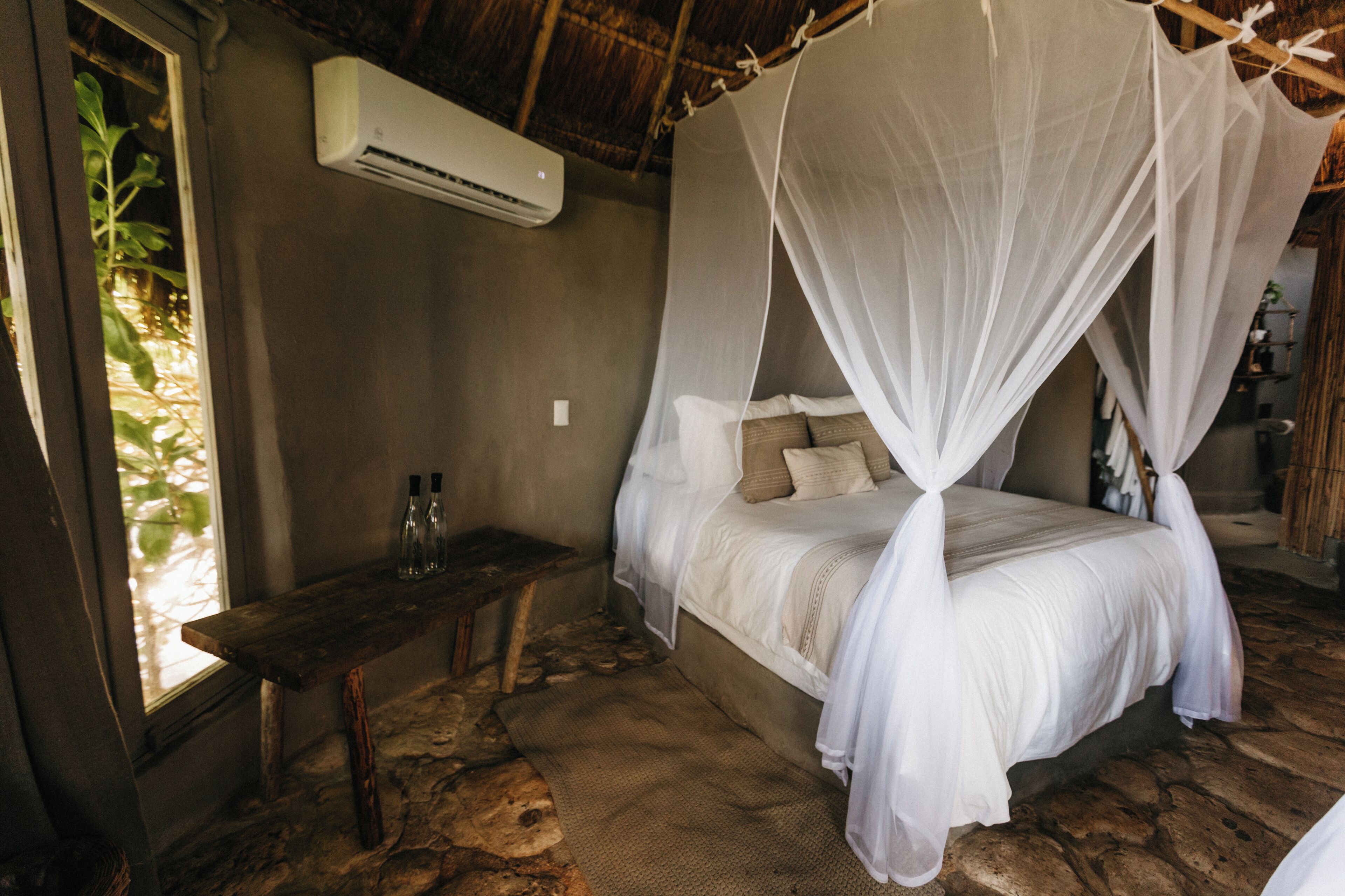 Een kamer met een bed onder een hemelbed, een bankje, een plank met flessen, en een airconditioning.