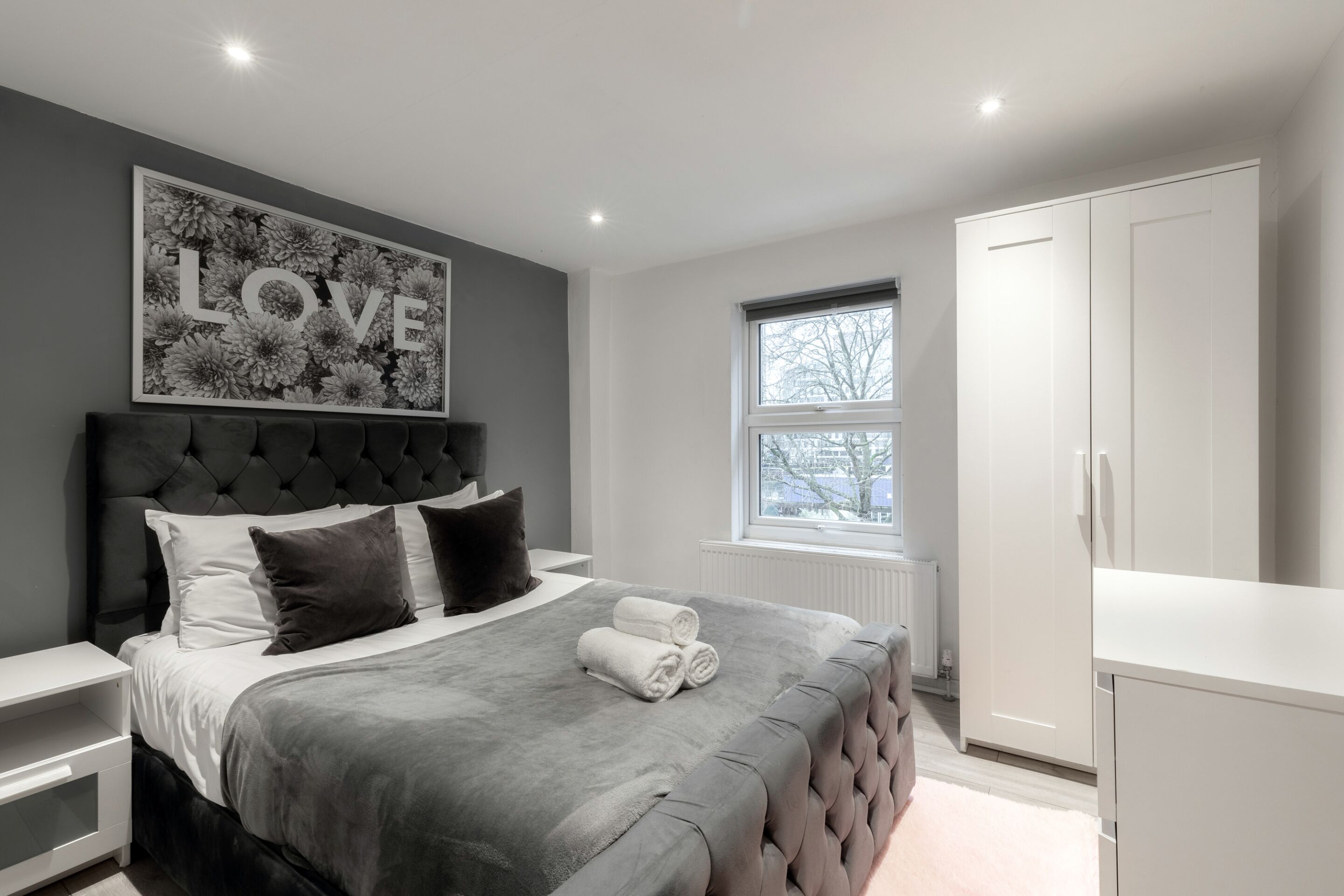 A modern bedroom with a tufted headboard, a large floral artwork, and neatly folded towels on the bed.