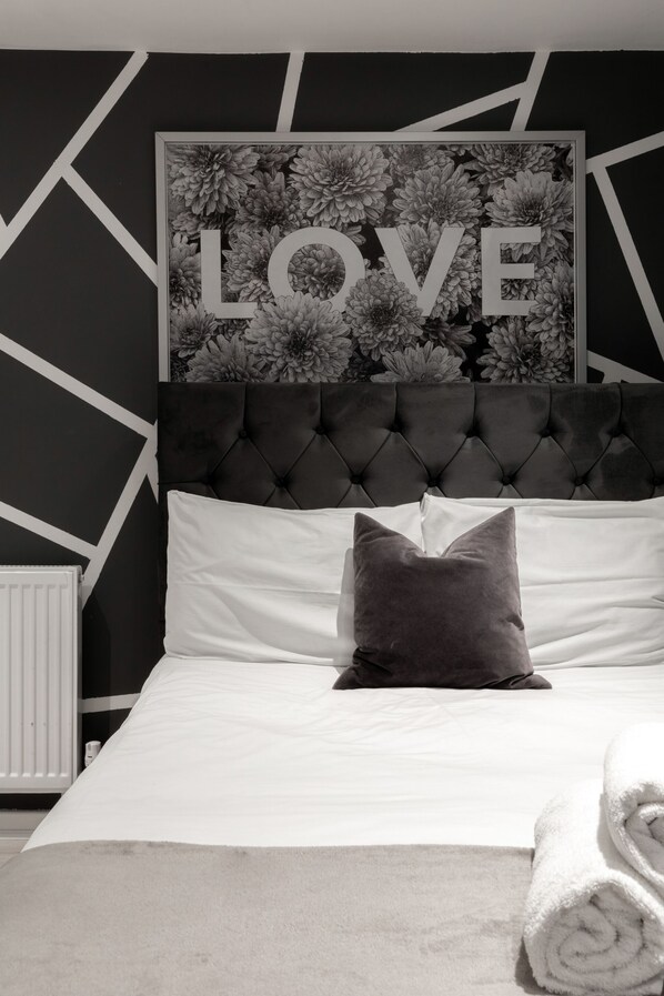 Un dormitorio con cabecera capitonada, ropa de cama blanca, una toalla enrollada y un cartel con la palabra 'LOVE' sobre la cama.