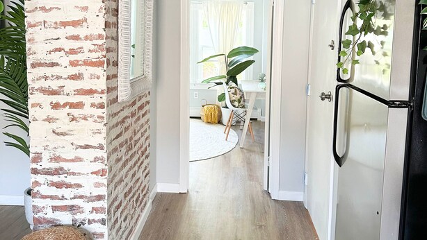 Un pasillo con pared de ladrillo, un otoman tejido y una puerta que da a una habitación con una planta.