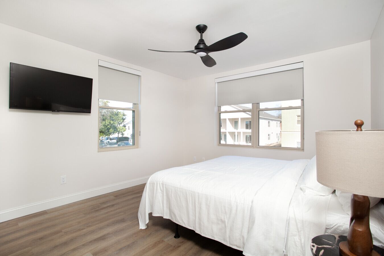 A bedroom with a bed, a ceiling fan, a TV mounted on the wall, and a window with a view of buildings outside.