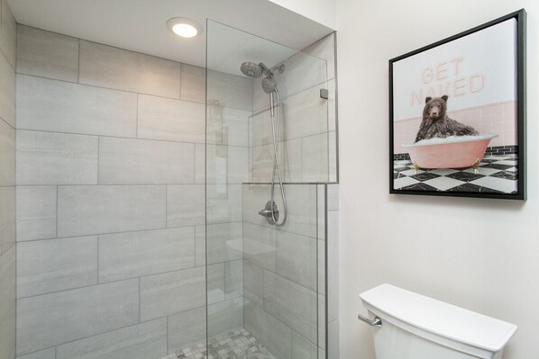 Een moderne badkamer met een douchecabine van glas, een wit toilet en een lijst met een foto van een beer in bad, met de tekst 'GET NAKED'.