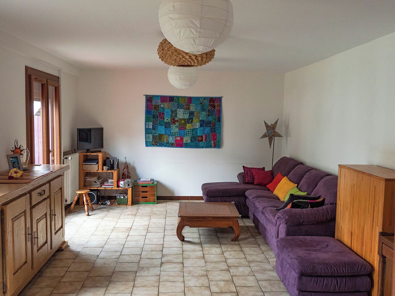 A living room with a purple sofa, a wooden coffee table, a television, and a colorful quilt hanging on the wall.