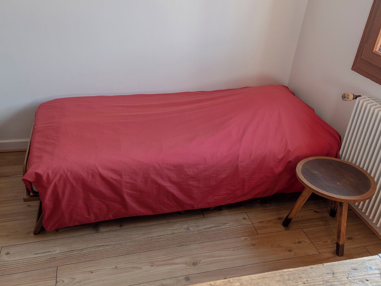 A small, single-bed room with a red bedspread, a wooden chair, and a radiator.