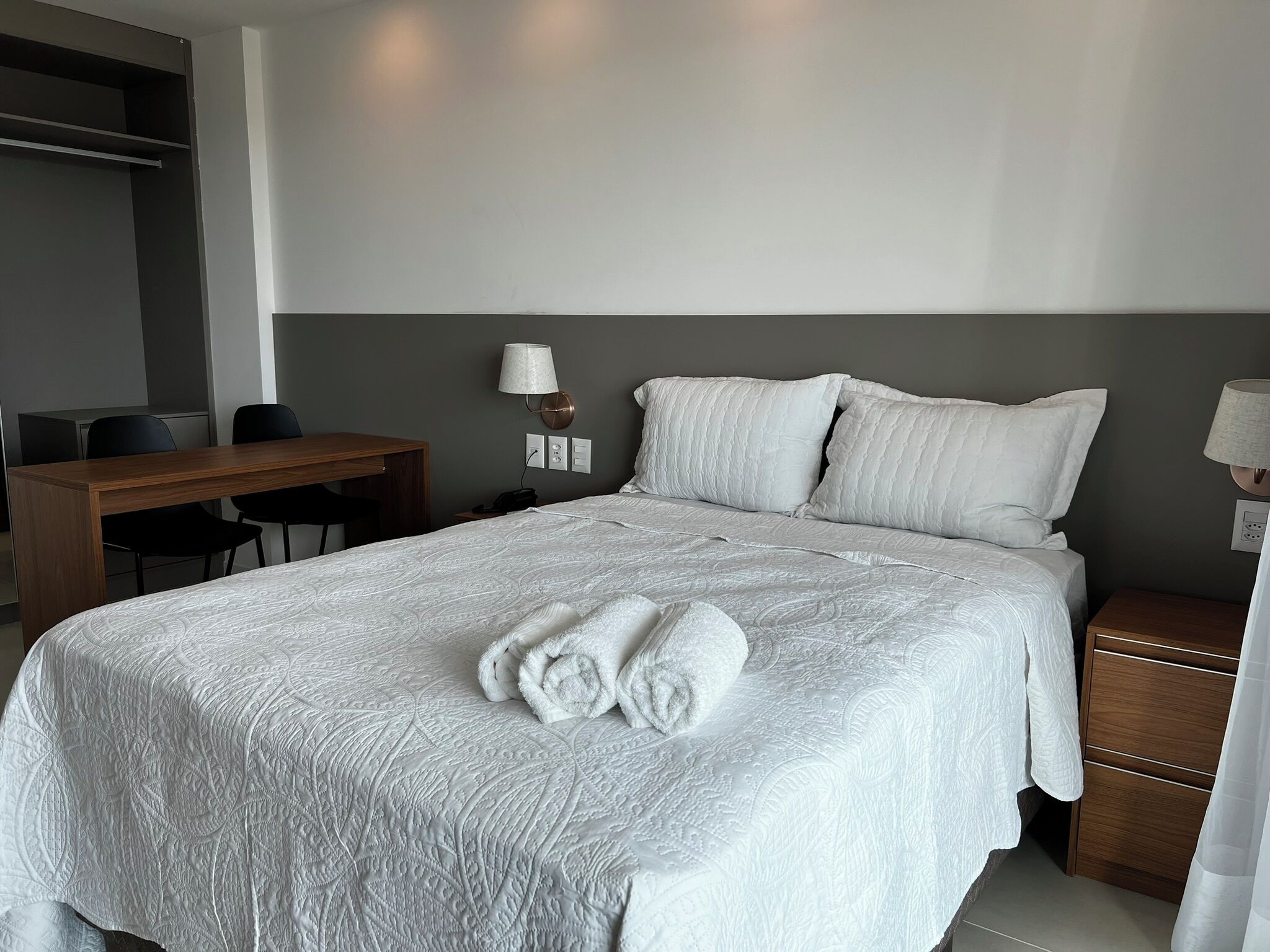 Ein ordentlich bezogenes Bett mit weißer Bettdecke und Kissen, ein Holztisch mit Stühlen und eine Wandlampe.