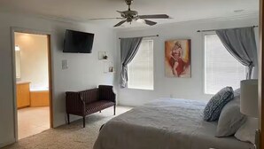 Un dormitorio con una cama, ventilador de techo, televisor montado en la pared y un cuadro.
