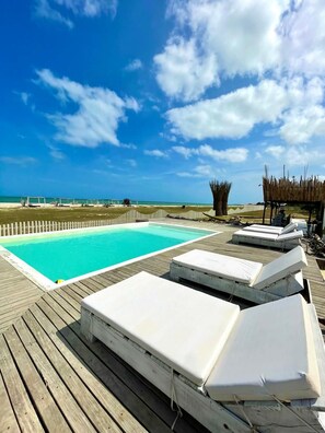 A wooden deck with a swimming pool, lounge chairs, and a view of the ocean. - Aiwa Hotel  (Manaure)