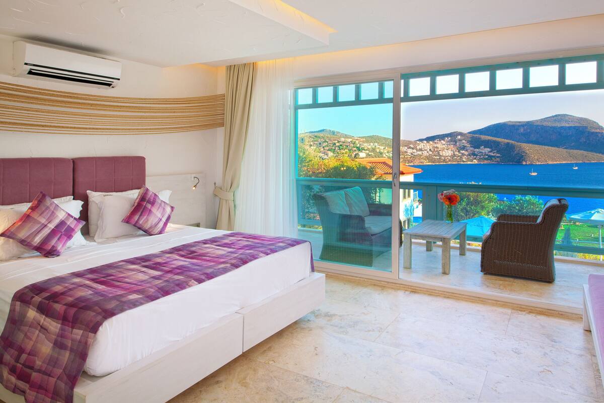A bedroom with a large bed, a balcony with a view of the sea and mountains, and a dining area with outdoor furniture.