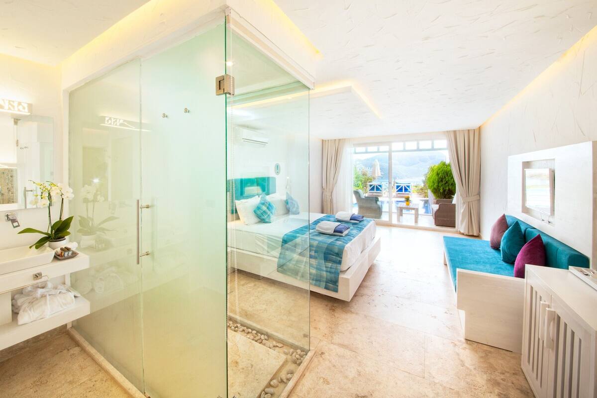 A modern hotel room with a glass-enclosed shower, a bed with a blue and white bedspread, a seating area with a sofa and armchairs, and a view of the outdoors.