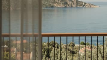 A view through a window with a balcony, overlooking a calm sea and a mountainous landscape.