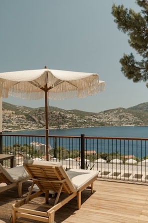 Une terrasse en bois avec deux chaises longues et un parasol blanc, offrant une vue sur un paysage côtier avec des collines et une étendue d’eau.
