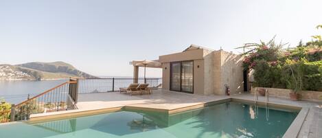 A swimming pool with a view of the sea, a covered outdoor seating area, and a stone building with glass doors.