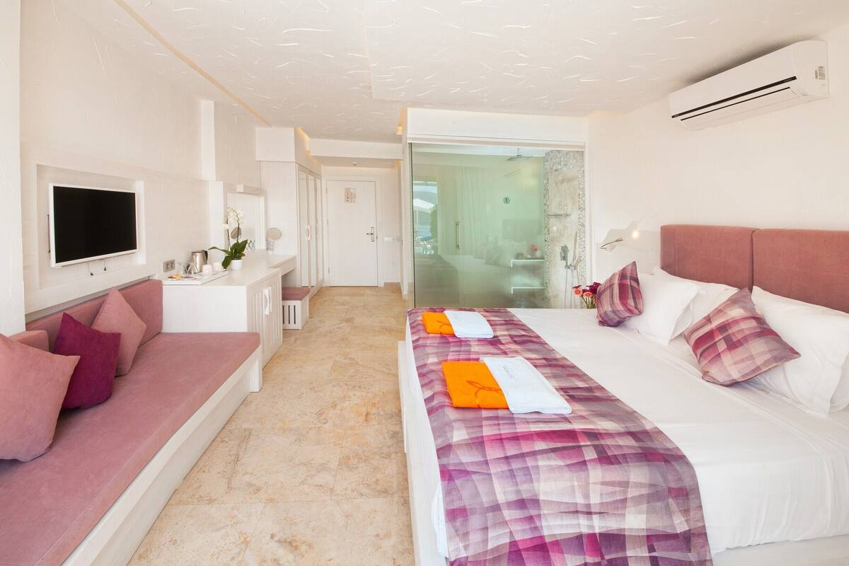 A modern hotel room with a bed, a sofa, a TV, and a bathroom visible through a glass door.