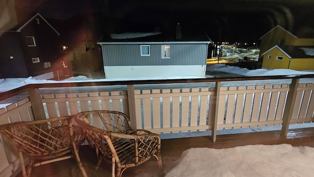 Un balcon avec une balustrade en bois, une chaise en osier et une vue sur des maisons se dressant au milieu d’un paysage enneigé.