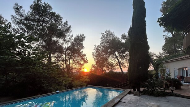 Une piscine avec vue sur le coucher de soleil, entourée d’arbres et bordée d’un bâtiment dont le toit est recouvert de tuiles.