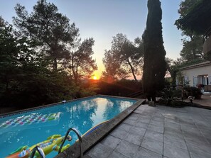A swimming pool with a view of the sunset, surrounded by trees and a building with a tiled roof.