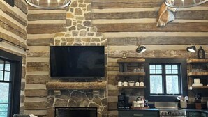 Un intérieur rustique avec une cheminée en pierre, des murs en bois, un téléviseur à écran plat, un coin repas et une cuisine équipée d’une cuisinière et d’une machine à café.