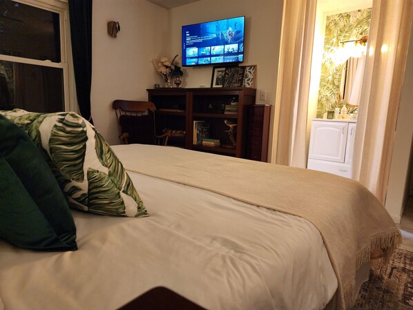 Una cama bien hecha con una colcha blanca y un almohada verde y blanca. Un televisor de pantalla plana está montado en la pared sobre un mueble de madera con libros y objetos decorativos.