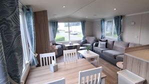 Ein modernes Wohnzimmer mit Essbereich, grauen Sofas, einem Holztisch und einem großen Fenster mit Vorhängen.